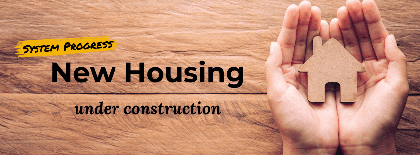 Photo of hands holding small house cut-out on wooden background with text that says "System Progress, new housing under construction"