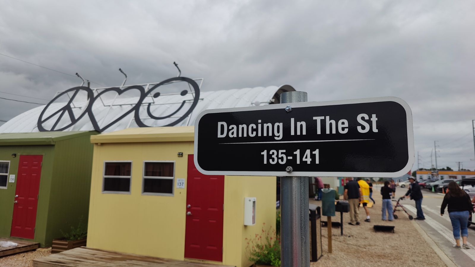 Street sign that says "Dancing in the St" with a tiny home in the background and peace, love, and happiness symbols